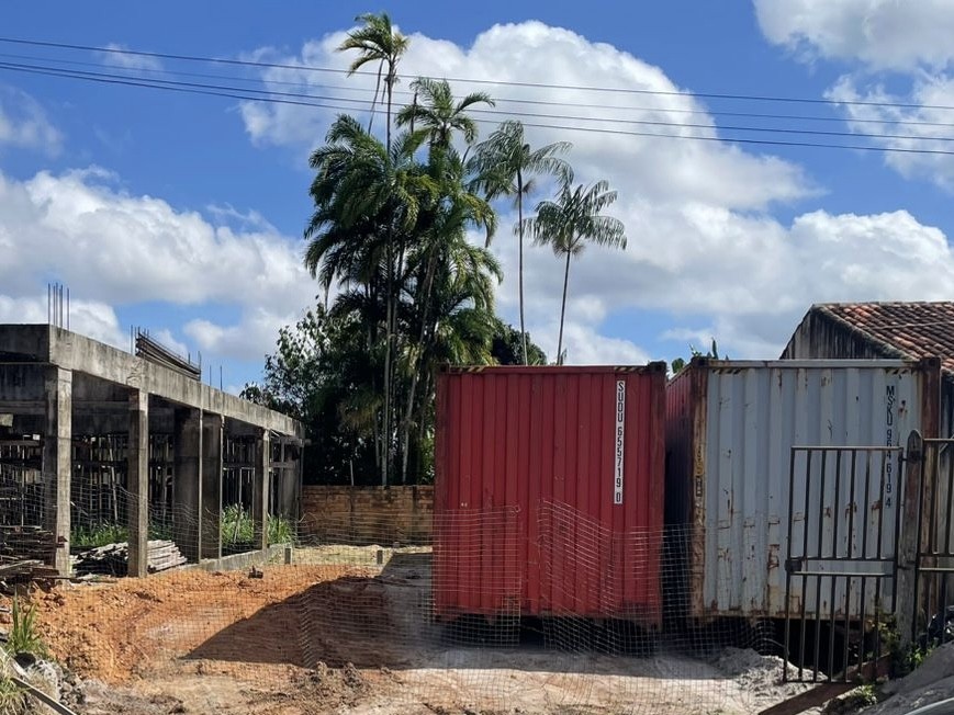 Canteiro de obras da Fast Escova com dois containers marítimos (um vermelho e um cinza) instalados em terreno de terra ao lado de uma estrutura de concreto em construção.