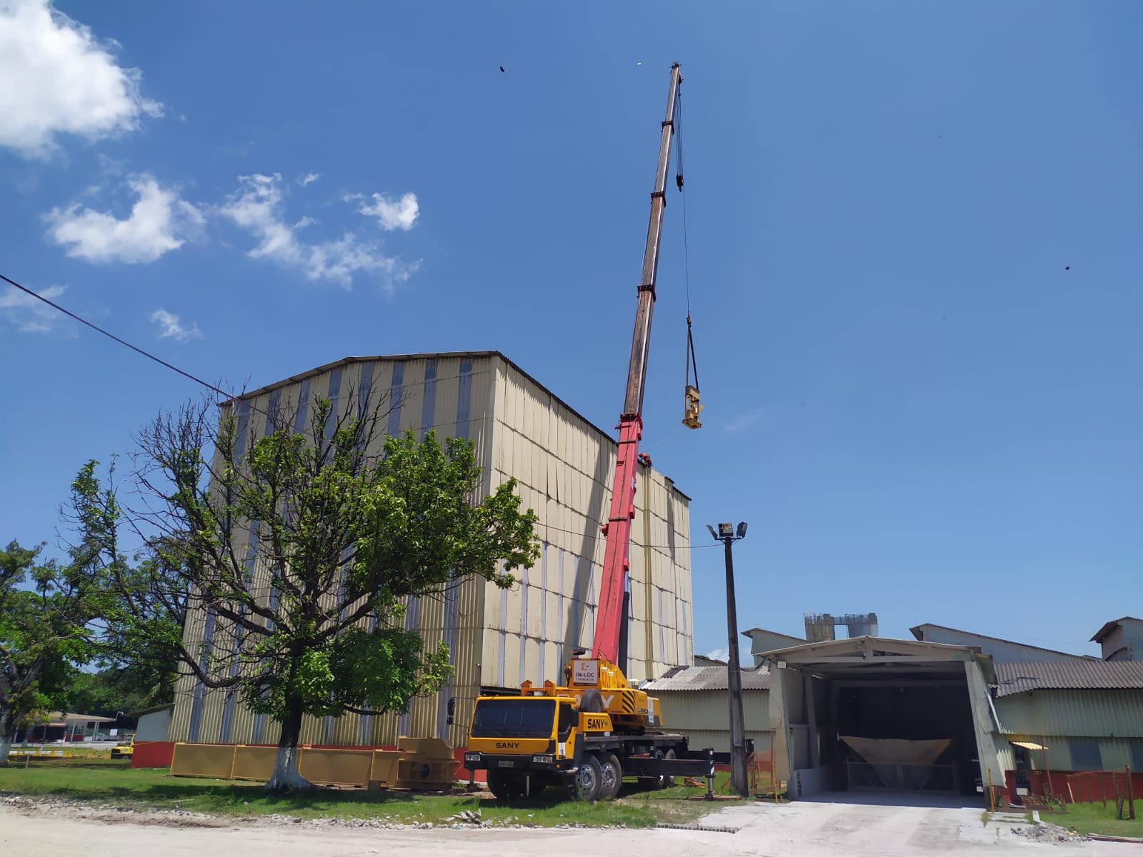 Caminhão guindaste Sany amarelo e vermelho realizando o içamento de carga pesada para montagem industrial em um galpão de grande porte sob céu limpo.