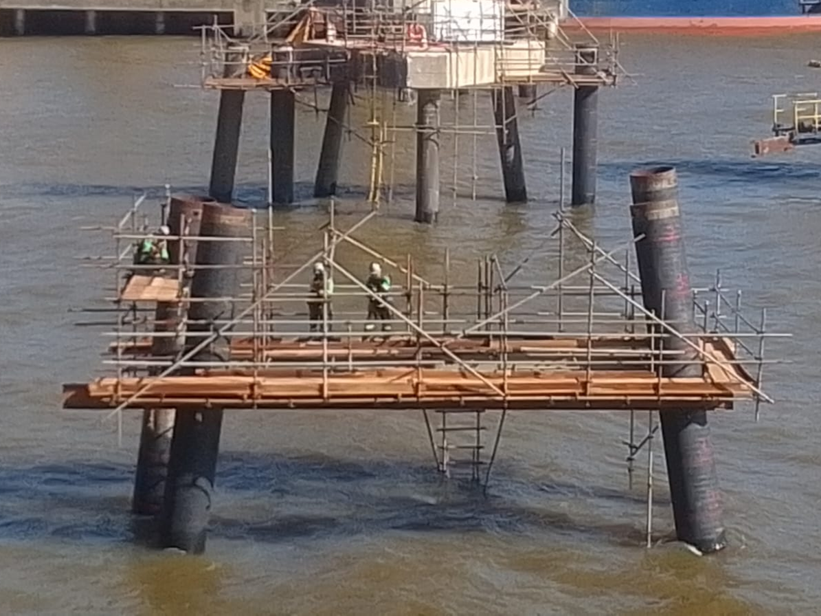 Operários em plataforma de andaime montada sobre estacas metálicas no rio, realizando manutenção estrutural em dolfim de atracação portuária em Barcarena.