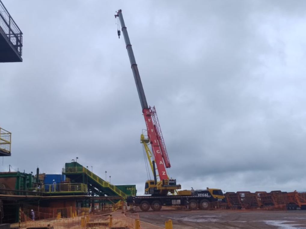 Guindaste telescópico da marca Eficaz com lança vermelha e preta estendida em ângulo acentuado, realizando operação de içamento em pátio industrial sob céu nublado.