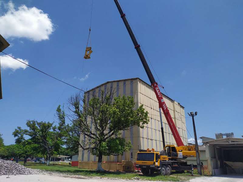 Guindaste articulado vermelho e amarelo operando o içamento de uma viga metálica próxima a um galpão industrial alto com revestimento metálico bege em Barcarena.
