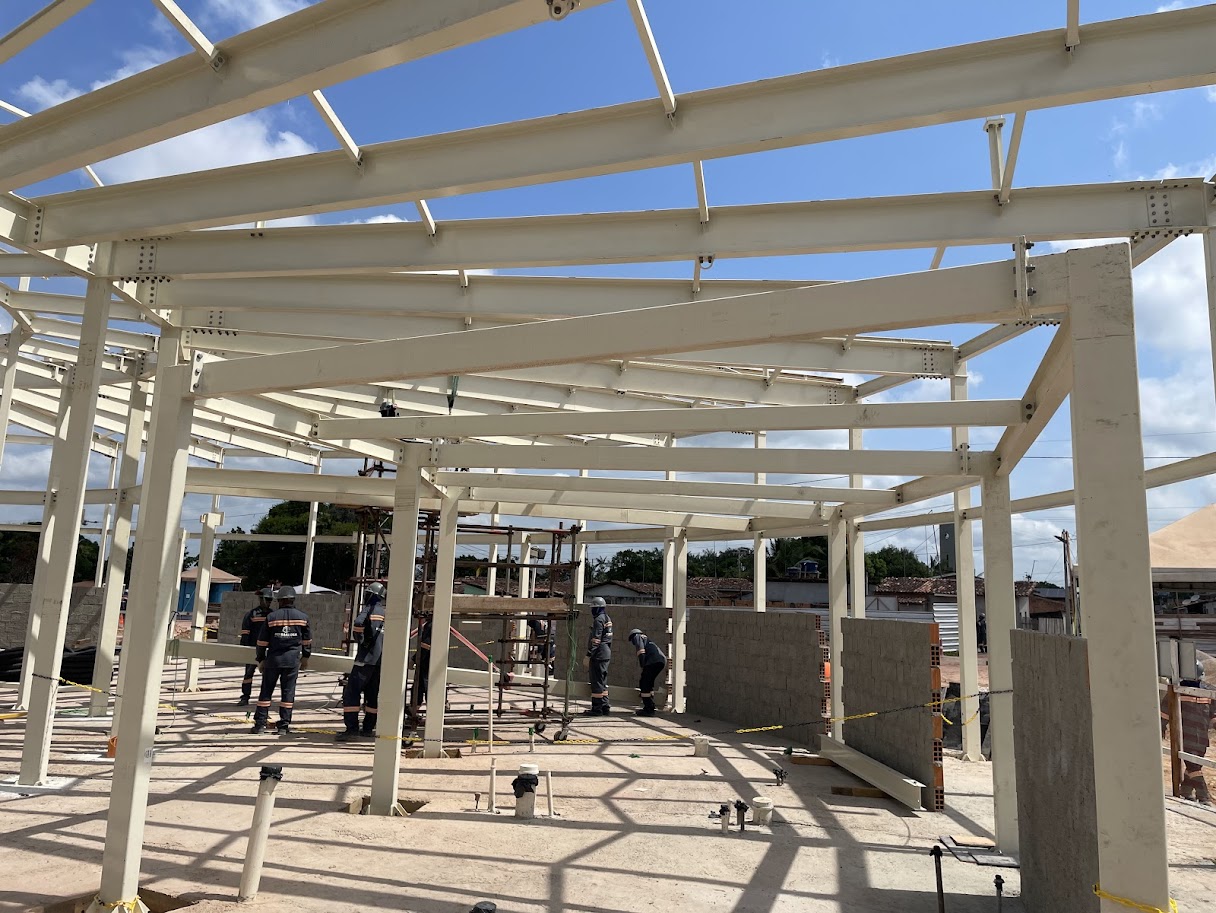 Vista interna de grande galpão em construção com estrutura metálica bege, operários uniformizados e paredes de bloco de concreto sendo levantadas em Moju.