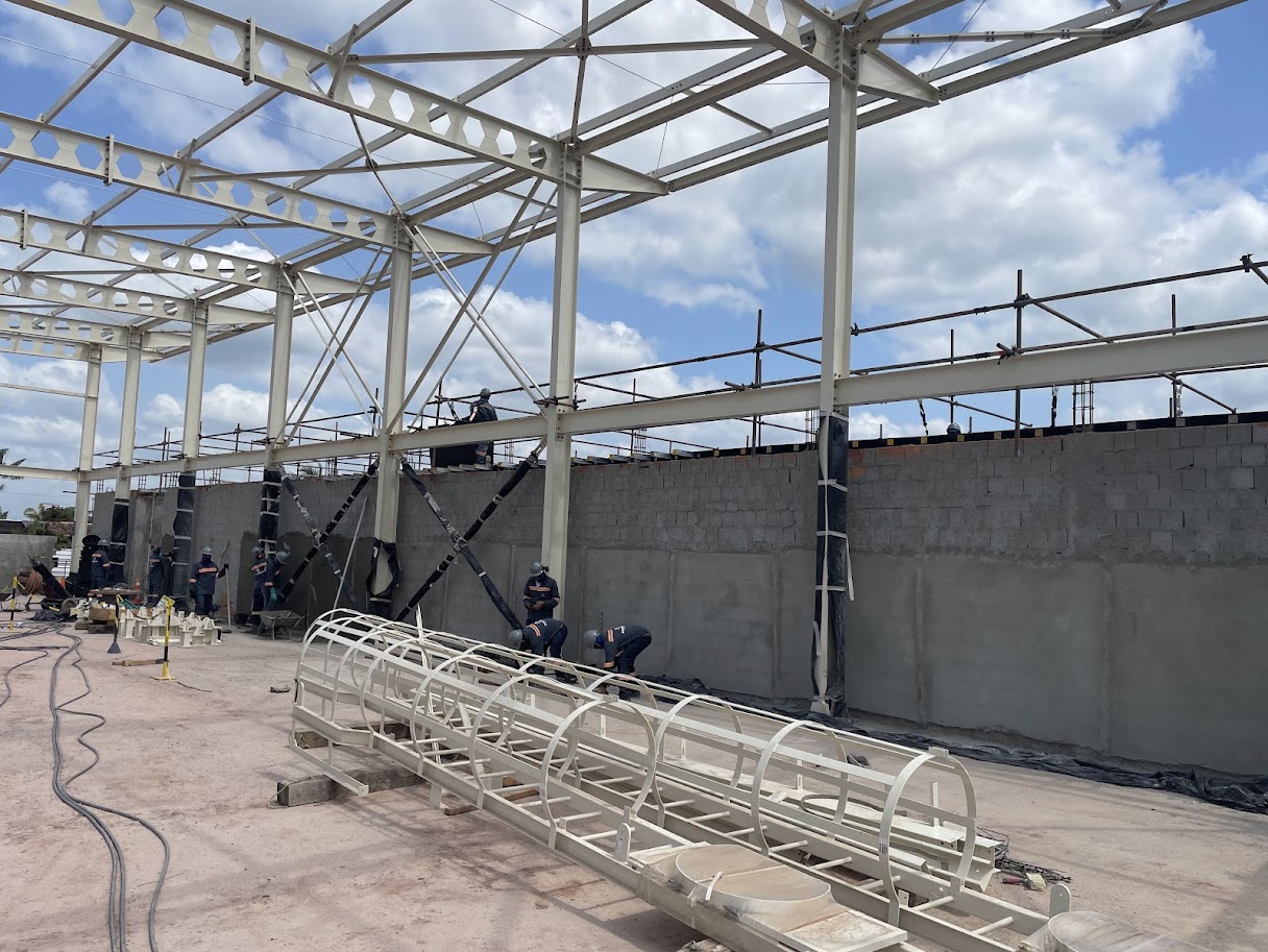 Canteiro de obras com estrutura metálica de galpão industrial, vigas alveolares beges e escada marinheiro com guarda-corpo metálico no chão em Barcarena.
