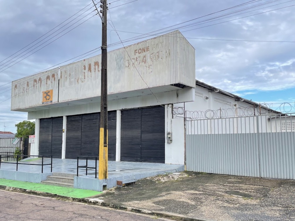 Fachada frontal de galpão comercial com testeira metálica branca, três grandes portões de aço pretos e rampa de acesso lateral protegida por cerca de arame.
