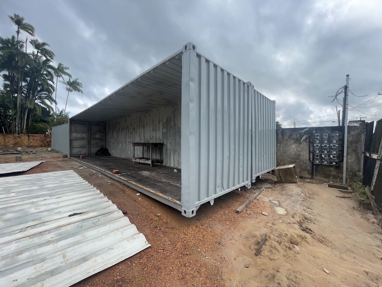 Container marítimo cinza aberto lateralmente em canteiro de obras, revelando o interior com piso de madeira compensada e bancada técnica ao fundo.