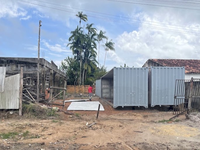 Canteiro de obras da Fast Escova com três containers cinzas posicionados em formato de "L" em um terreno preparado, com operário ao fundo.