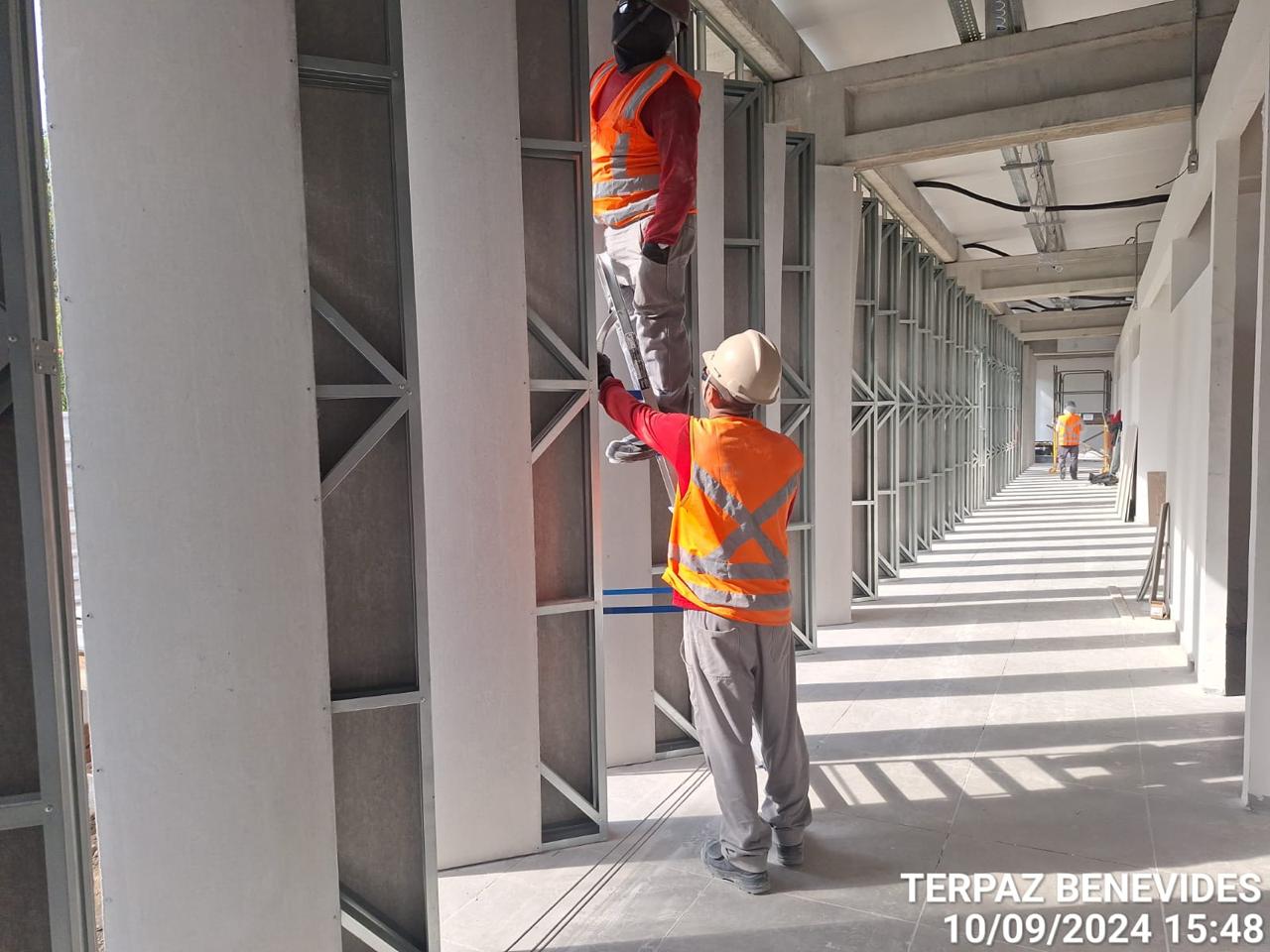 Dois operários com coletes reflexivos laranja e capacetes instalando divisórias de drywall em um corredor longo de um prédio em construção (Terpaz Benevides).
