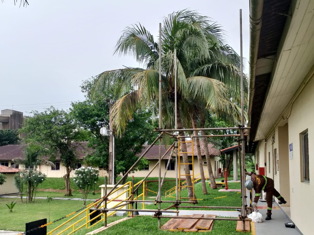 Laudos Mecânicos – F.G. Construção Operário com uniforme marrom e capacete cinza montando estrutura de andaime tubular externo em área gramada próxima a palmeiras e prédios administrativos.