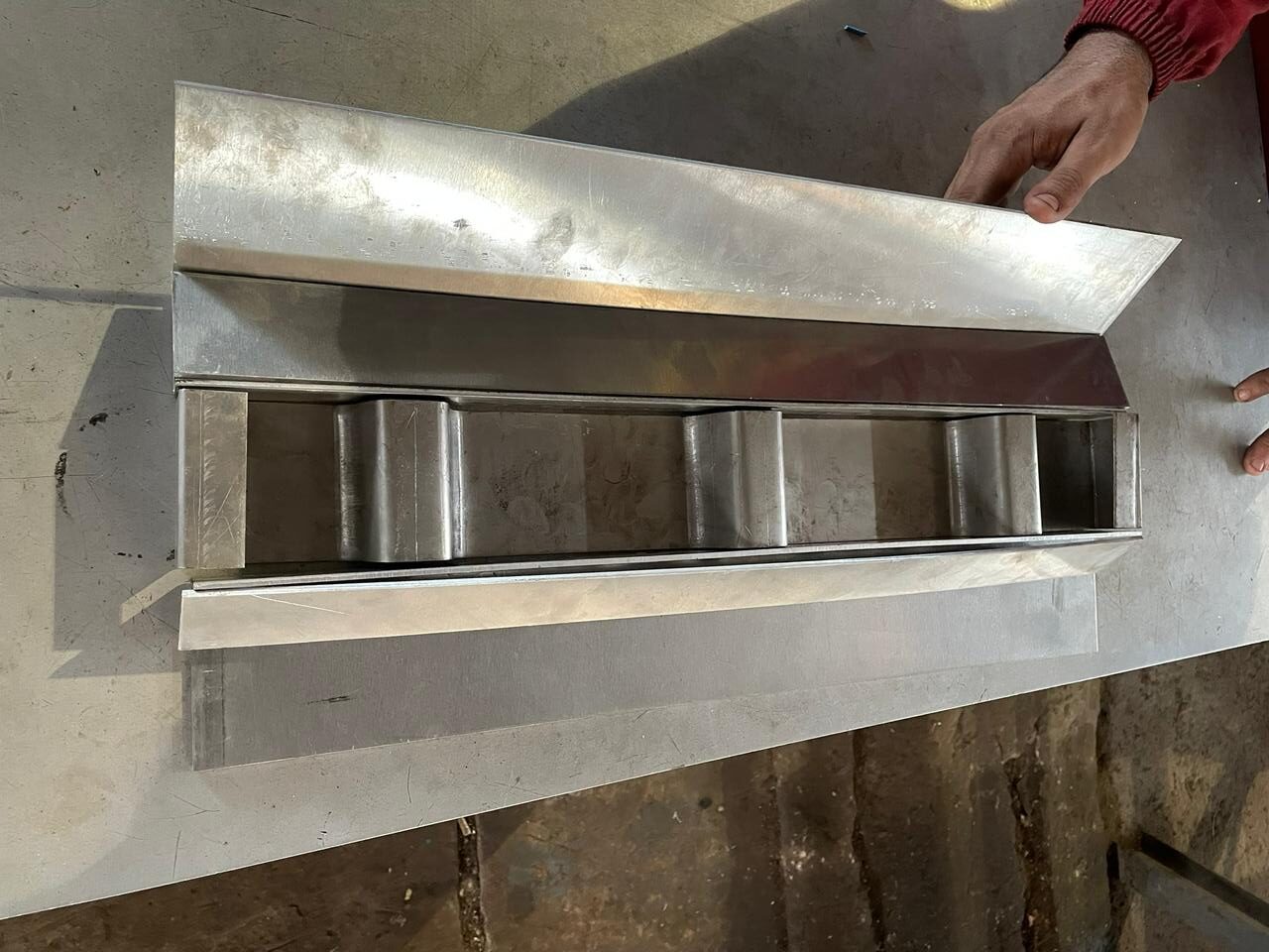 Detalhe de calha industrial metálica em aço inox com divisórias internas soldadas e abas laterais para fixação, sendo apresentada em bancada de oficina.