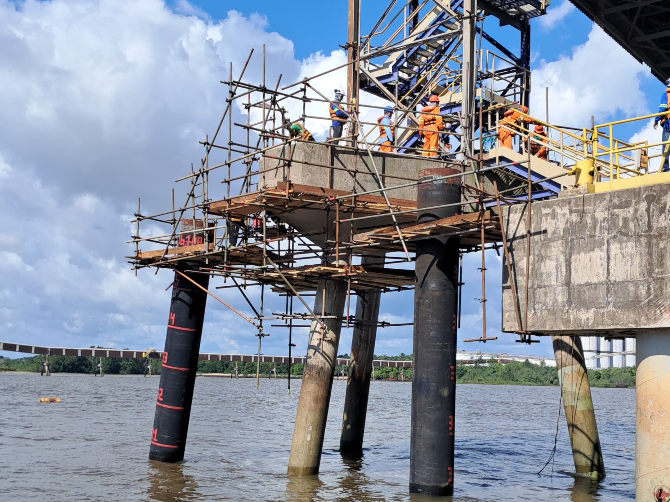 Estrutura de andaimes tubulares montada sobre dolfins de concreto e estacas metálicas no Terminal de Grãos Ponta da Montanha (TGPM), com operários em plataforma elevada sobre o rio.