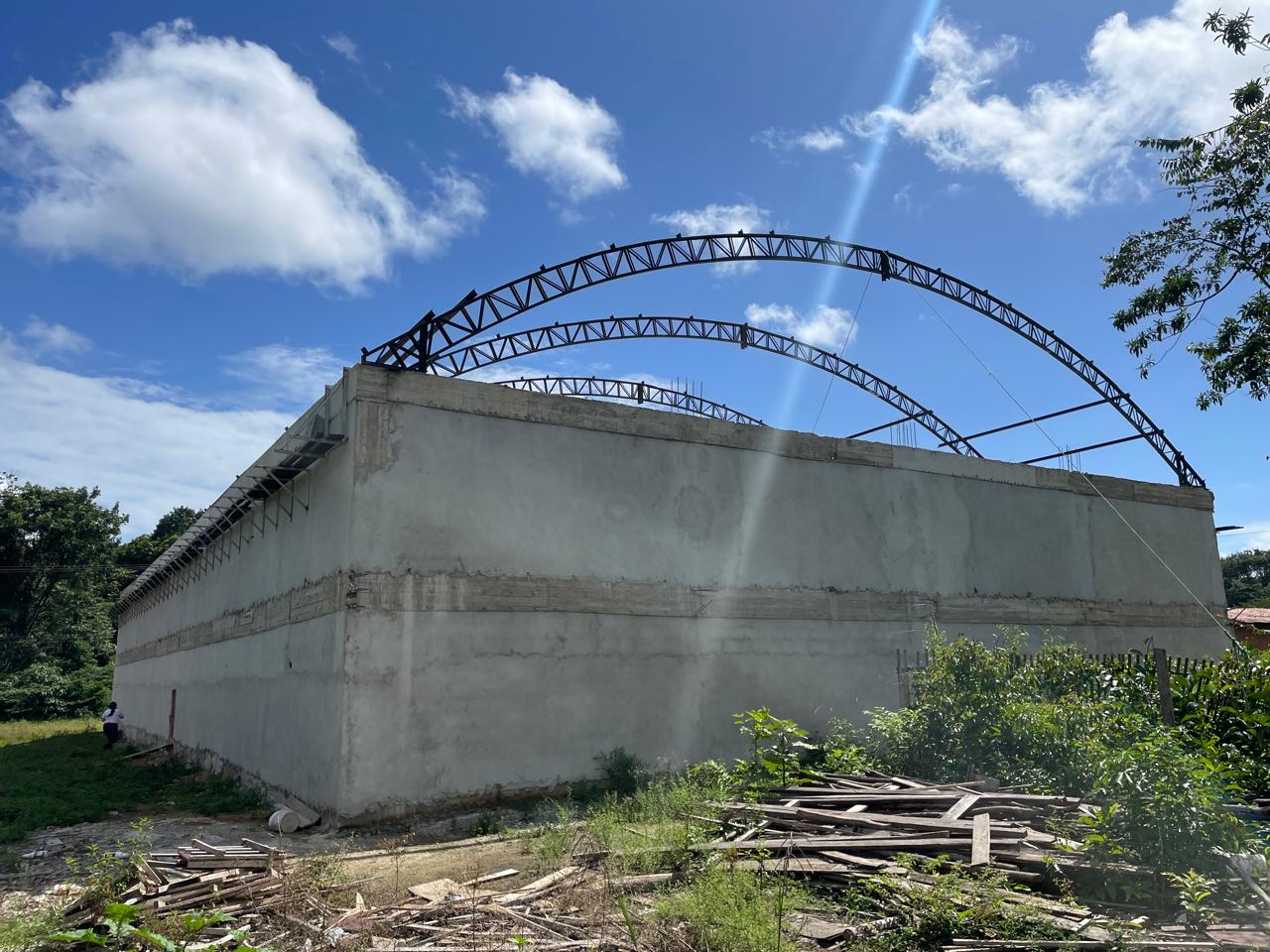 Vista lateral de grande galpão comercial com paredes de concreto e início da montagem de arcos metálicos treliçados para cobertura sob céu azul.