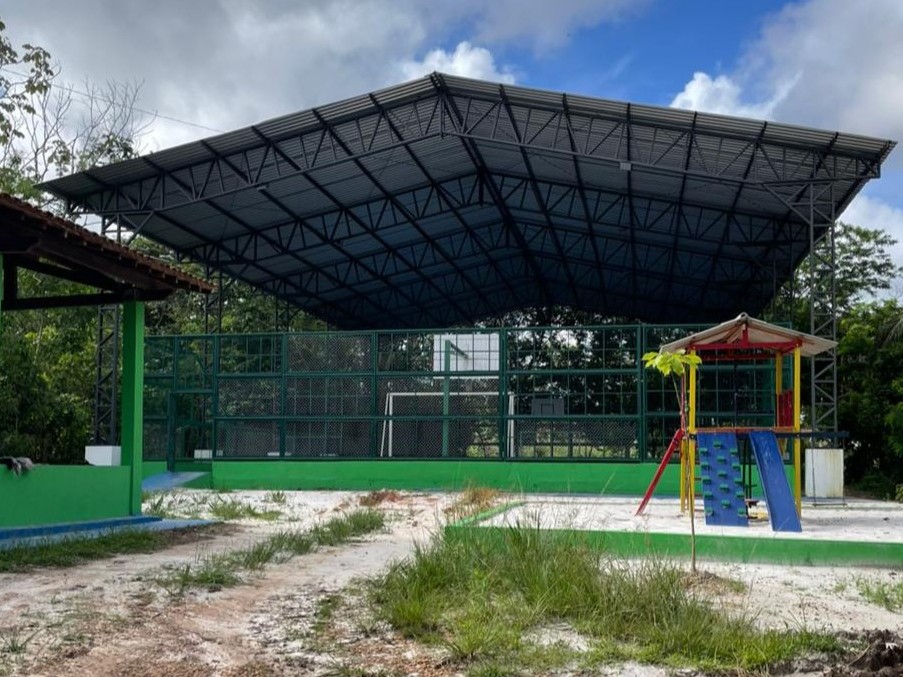 Quadra poliesportiva com cobertura metálica cinza, fechamento lateral em tela verde, traves de futebol e parquinho infantil colorido ao lado em Barcarena.