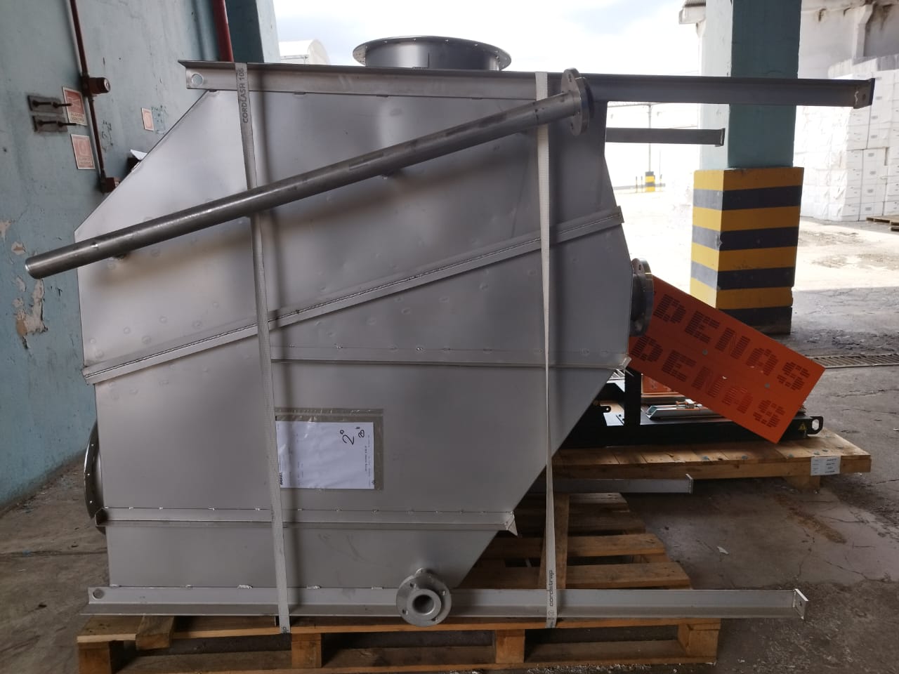 Tanque industrial metálico de grande porte acomodado sobre palete de madeira em área de expedição, com cintas de amarração para transporte.