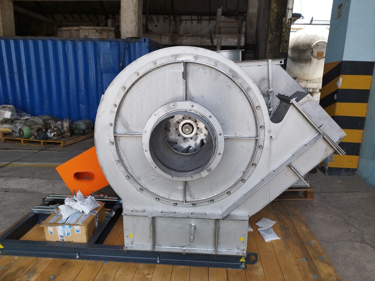 Ventilador centrífugo industrial de grande porte em aço inox, montado sobre base metálica e palete de madeira em área de estoque.
