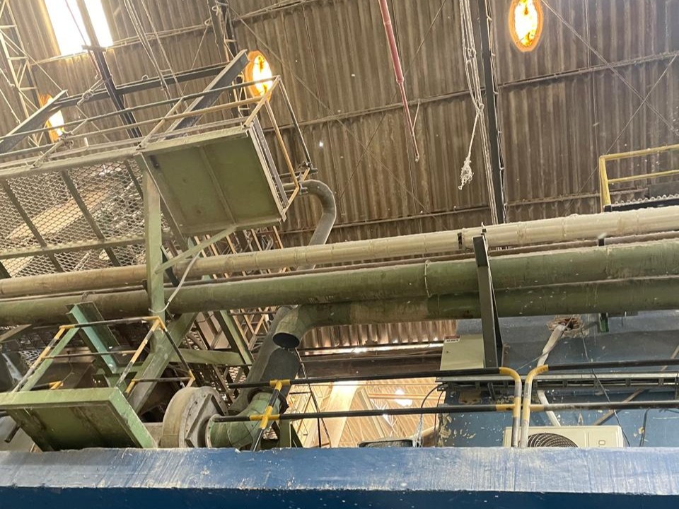 Vista inferior de galpão industrial com telhado de fibrocimento, tubulações de exaustão horizontais e passarelas metálicas de manutenção suspensas.