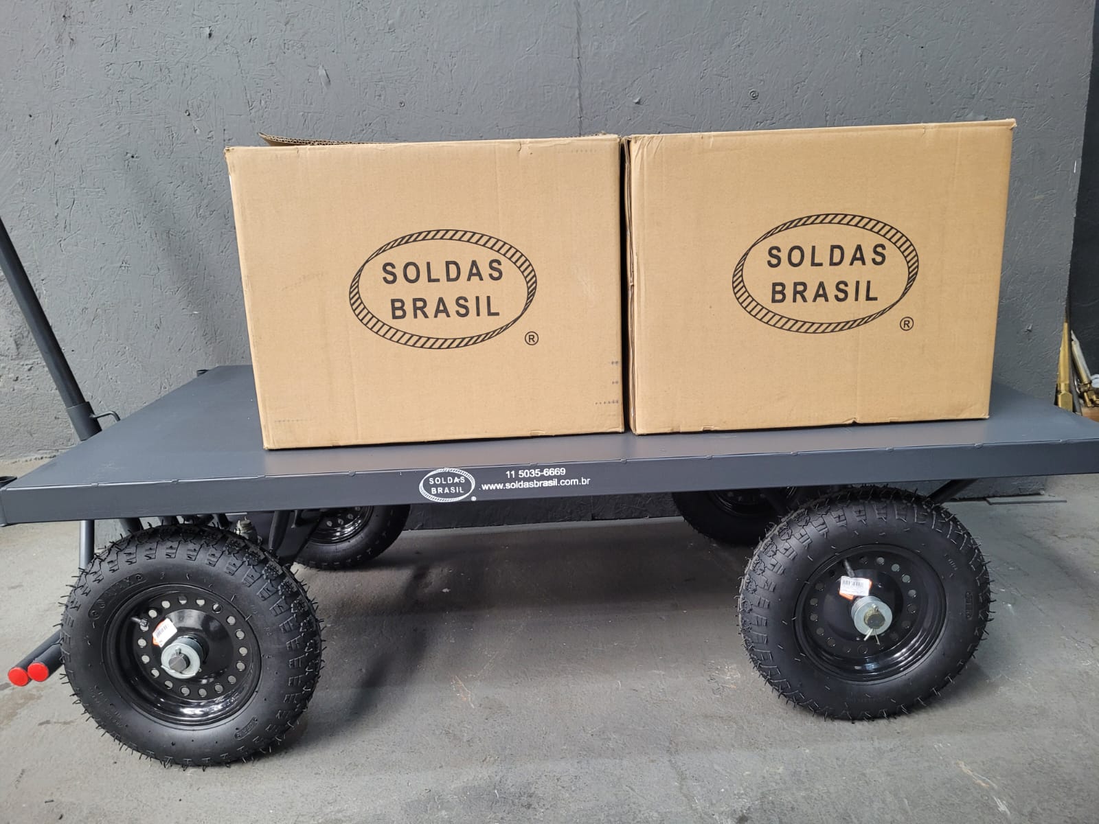 Carrinho de carga industrial cinza com quatro rodas pneumáticas pretas e duas caixas de papelão da marca "Soldas Brasil" em cima da plataforma.