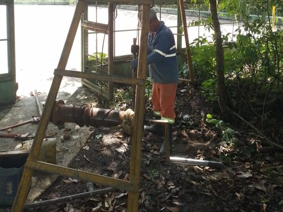 Operário de uniforme laranja e azul realizando sondagem de solo a percussão (SPT) com tripé metálico em área de mata para o projeto da Quadra Burajuba.