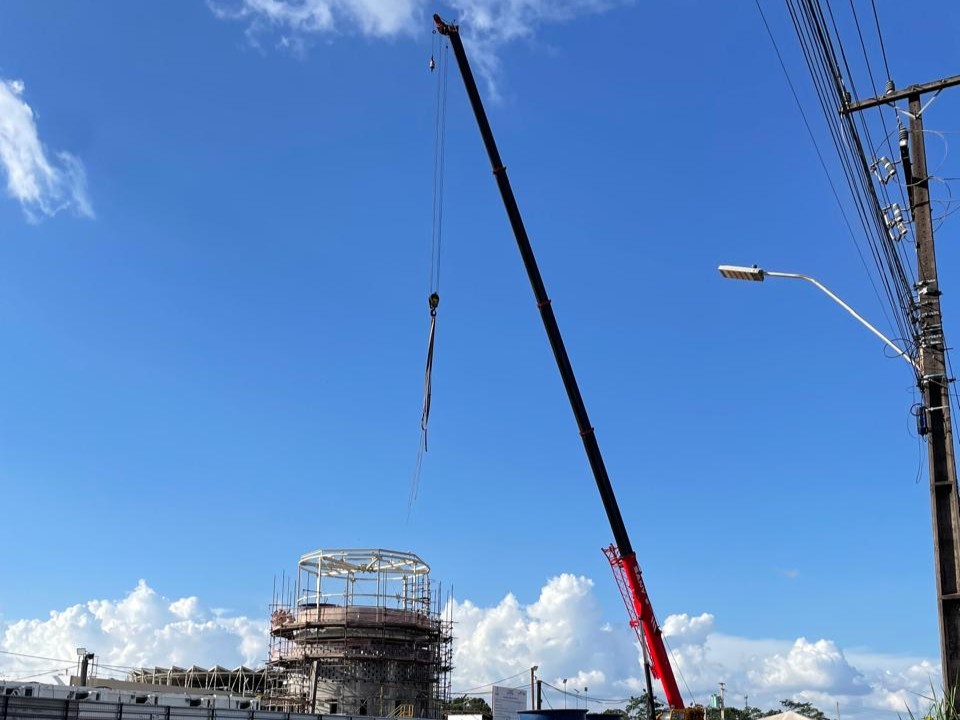 Guindaste vermelho com lança telescópica estendida realizando o içamento de componentes metálicos para a montagem de um grande tanque industrial circular com andaimes.