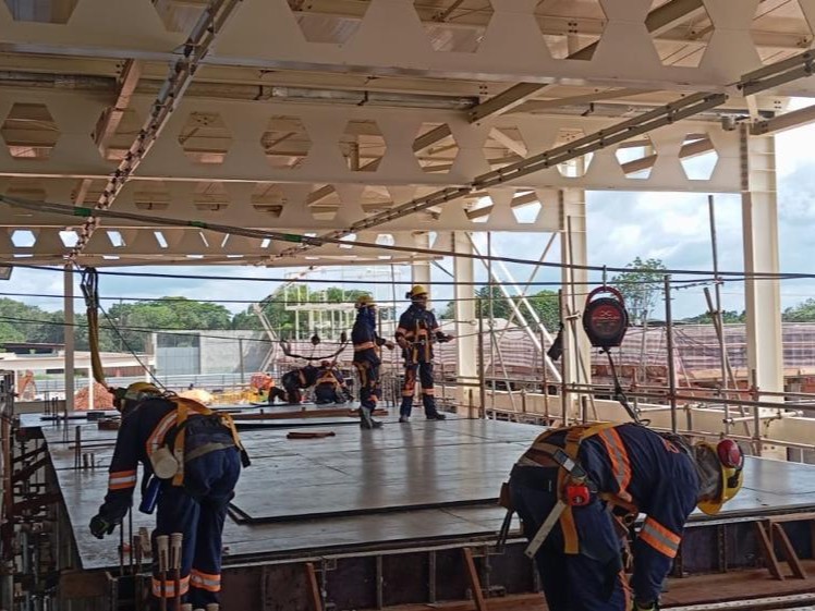 Equipe de operários com uniformes de alta visibilidade e cinturões de segurança trabalhando na montagem de laje sob estrutura metálica com vigas alveolares.