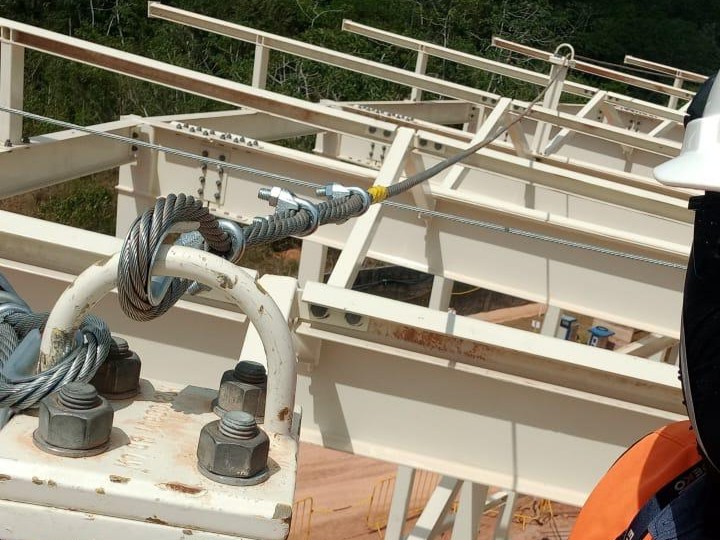 Detalhe técnico de ancoragem de linha de vida com cabo de aço, sapatilhas e grampos fixados em estrutura metálica robusta com parafusos de alta resistência.