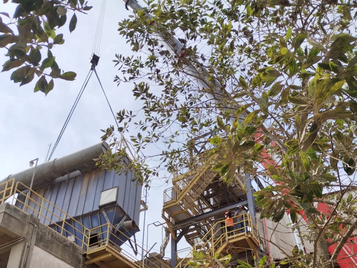 Laudos Mecânicos – F.G. Construção Vista inferior de guindaste realizando o içamento de uma tubulação metálica de grande diâmetro conectada a um filtro de mangas azul, com operários observando de uma plataforma metálica elevada.