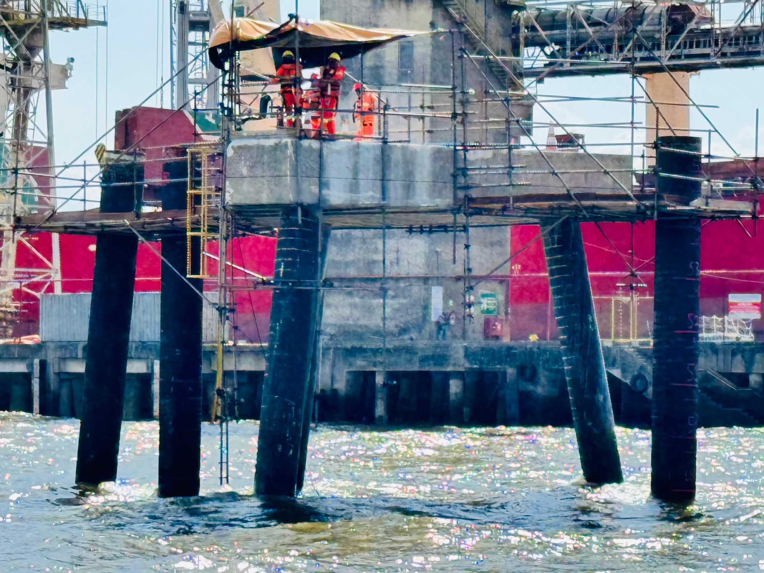 Equipe de operários em plataforma elevada sobre estacas metálicas no mar, realizando manutenção em dolfim de atracação com apoio de andaimes e cobertura de proteção.