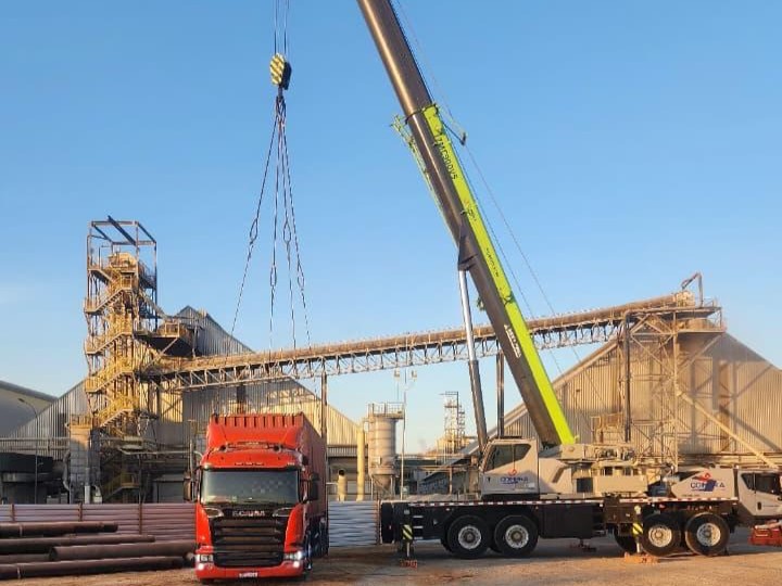 Guindaste Zoomlion branco e verde realizando içamento de carga sobre caminhão Scania vermelho, em frente a uma estrutura de esteira transportadora e galpões industriais da Coimbra.